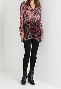 Bluse mit Blumendruck, langen Ärmeln in Pink und Schwarz, mit tiefem V-Ausschnitt und fließender Silhouette, kombiniert mit schwarzen, enganliegenden Hosen und Stiefeletten.