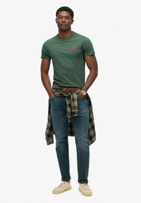 Grøn bomulds t-shirt med rødt logo, blå denim jeans og beige sneakers. Mørkternet skjorte bundet om taljen. Afslappet pasform og rene linjer.