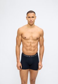 Γυμνασμένος άνδρας που στέκεται κατά πρόσωπο, φοράει μποξεράκια Tommy Hilfiger σε ναυτικό μπλε χρώμα με λογότυπο της μάρκας στη μέση, σε απλό λευκό φόντο.