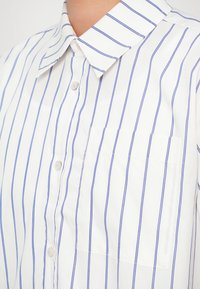 Bela srajca s gumbi in navpičnimi modrimi črtami, s ovratnikom in žepom na prsih. Tkanina se zdi gladka in lahka.