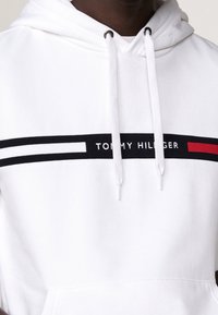 Felpa bianca con striscia orizzontale nera e rossa. Logo "TOMMY HILFIGER" in nero all'interno della striscia. Coulisse e texture del tessuto morbida.