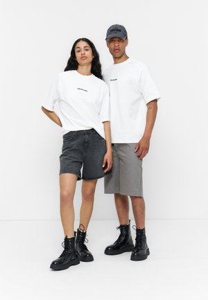 Νέος άνδρας και γυναίκα που φορούν λευκά t-shirts «YourTurn», σορτς και μαύρα μπότες, στέκονται μπροστά σε ένα απλό λευκό φόντο.