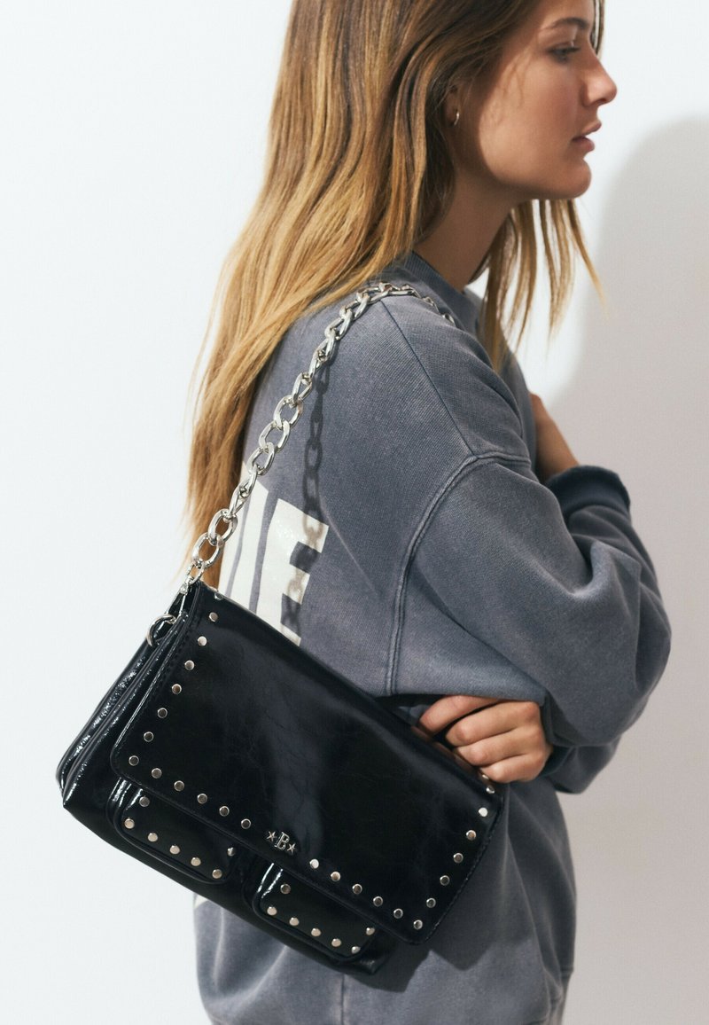 Jeune femme en sweat-shirt gris porte un sac à épaule en cuir noir clouté avec une bandoulière en chaîne argentée sur fond blanc.