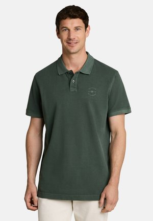 Man met kort donker haar, die een donkergroen polo met korte mouwen en lichte broek draagt, staand tegen een effen achtergrond.