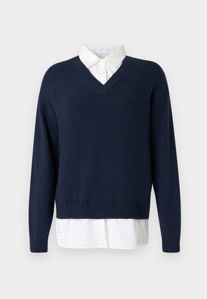 Suéter de punto suave en azul marino con escote en V, superpuesto a una camisa blanca con pequeños lunares esparcidos y un cuello clásico.