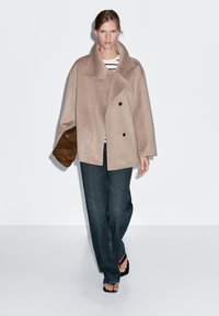 Beige vlněný kabát s dvojitým zapínáním, velkým límcem a oversize střihem, nošený přes pruhovaný top a tmavé džínové kalhoty.