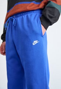 Modré tepláky Nike s elastickým pasem a šňůrkou. Mají boční kapsy a bílé logo Nike na levém stehně. Měkká textura látky.