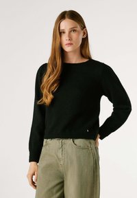 Melna adīta džemperis ar apaļu kakla izgriezumu un garām piedurknēm, valkāts kopā ar gaiši zaļām biksēm. Apakšmalā ir nemanāms logo.