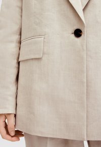 Blazer in lino beige con un singolo bottone rotondo, una tasca a pattina sul lato sinistro e una linea dritta. Texture liscia e design minimalista.