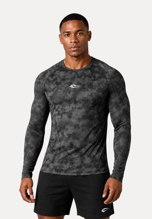 Uomo atletico che indossa una maglia mimetica nera e grigia a maniche lunghe e pantaloncini neri con logo bianco, in piedi su uno sfondo semplice.