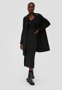 Schwarzer langer Mantel über einem figurbetonten schwarzen Kleid mit strukturiertem Oberteil und schlanker Silhouette, kombiniert mit schwarzen Knöchelstiefeln.