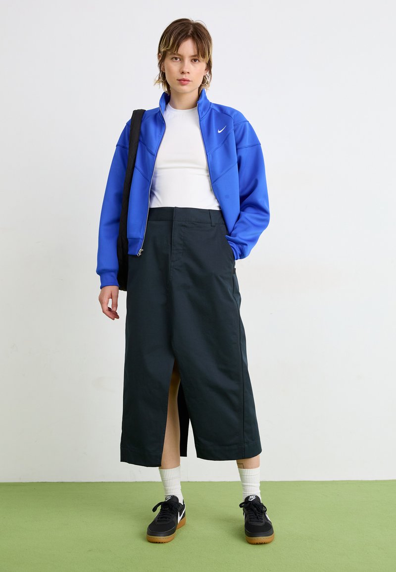 Veste zippée bleue, t-shirt blanc ajusté, jupe mi-longue vert foncé avec fente latérale, baskets noires et chaussettes blanches. Textures et couleurs unies.