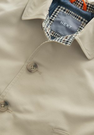 Beige trenchcoat med dubbelknäppning, med ett rutigt foder i blått, brunt och vitt, samt runda knappar. Lappen synlig.