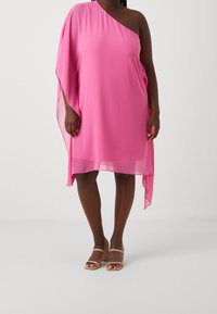 Femme portant une robe asymétrique rose à une épaule avec un tissu transparent et des sandales à talons beiges, se tenant sur un fond blanc.