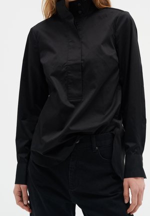 Femme portant une chemise noire à manches longues boutonnée, rentrée dans un pantalon noir, debout devant un fond clair uni.