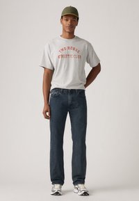 Grijze T-shirt met korte mouwen met "TWO HORSE ATHLETIC CLUB" in rode opdruk, gecombineerd met donkerblauwe jeans en grijs-zilveren sportschoenen.
