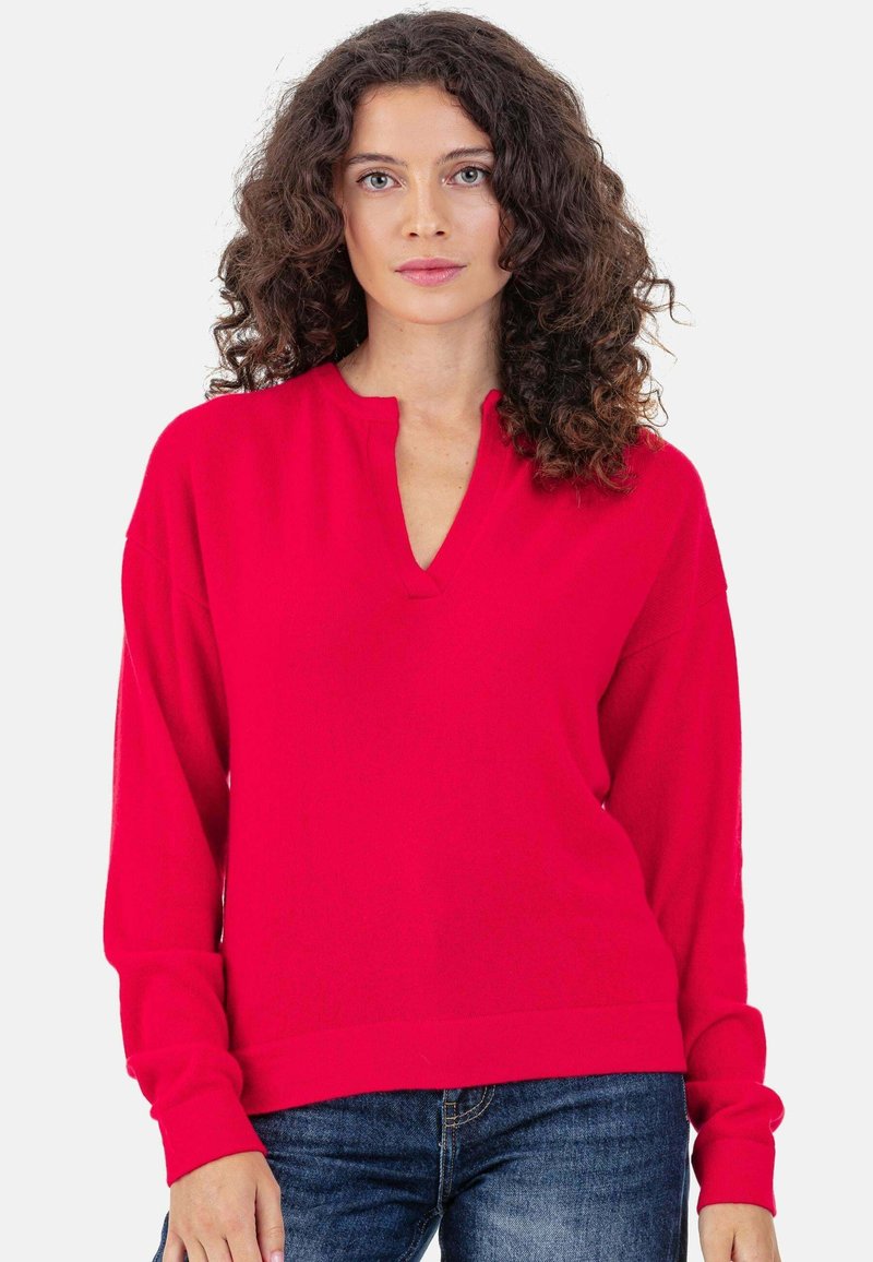 Pull rouge en maille avec un col en V, des manches longues et des poignets côtelés. Texture douce et coupe légèrement ample, associé à un jean bleu.