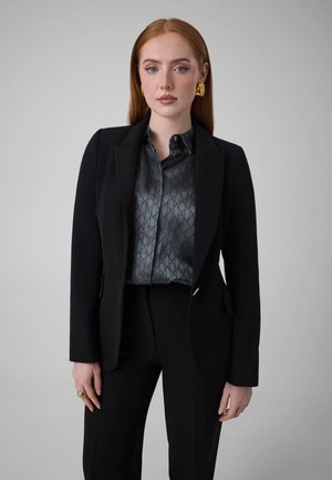 Mujer con el cabello largo y rojo lleva un traje negro, blusa gris con estampado, pendientes de oro y un anillo, de pie frente a un fondo gris liso.