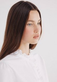 Biała bluzka z falbanistym kołnierzykiem i zapięciem na guziki, z długimi rękawami i gładką fakturą materiału. Modelka z prostymi, brązowymi włosami.