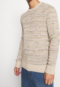 Maglione lavorato a maglia beige con collo a giro, caratterizzato da sottili variazioni di colore e polsini e orlo a coste, indossato sopra jeans blu.