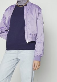 Osoba oblečená v světle fialové bomberce přes námořnický svetr, svetle modrých džínách, zlatém řetízku a světle šedém hidžábu na jednobarevném pozadí.