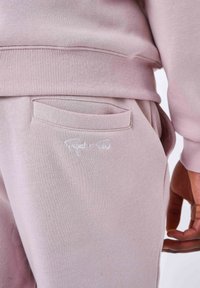 Pantalones de chándal rosa claro con un bolsillo trasero que tiene una solapa curva sutil, y un logo bordado en blanco "Project x Paris" en el costado.
