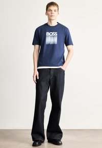Temno modra bombažna majica s prelivajočim logotipom "BOSS", združena z ohlapnimi temnimi jeansi in črnimi čevlji. Preprost dizajn in sodoben kroj.
