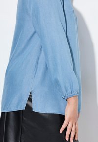 Camicia di jeans azzurro chiaro con maniche lunghe arricciate e spacchi laterali, caratterizzata da una texture liscia e una vestibilità rilassata.