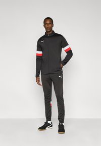 Μαύρο αθλητικό σετ με jacket με φερμουάρ, με κόκκινες και λευκές λωρίδες στα μανίκια και παντελόνι που ταιριάζει. Μαύρα αθλητικά παπούτσια ολοκληρώνουν την εμφάνιση.