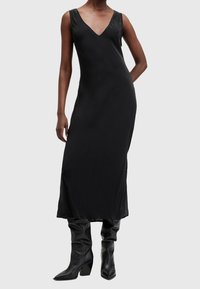 Robe maxi noire sans manches avec un décolleté en V, texture lisse et coupe légèrement décontractée. Portée avec des bottes noires à hauteur de genoux, décontractées.