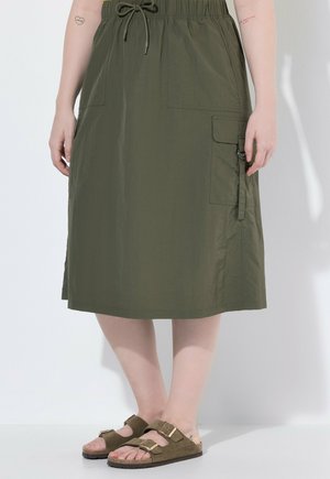 Femme portant une jupe mi-longue vert olive avec des poches et des sandales vert olive à boucle, debout devant un fond clair uni.