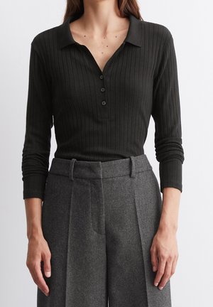 Femme portant un polo noir côtelé à manches longues rentré dans un pantalon en laine gris taille haute, coupe ajustée.