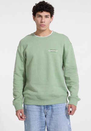 Lichtgroene sweatshirt met een ronde halslijn en mouwen met manchet. Bevat het logo "GUESS JEANS" in het wit op de borst. Gedragen met versleten blauwe jeans.