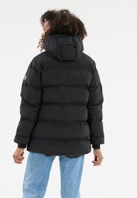 Person med krøllet hår iført en sort hættetrøje og lyseblå jeans, vendt væk mod en ensfarvet hvid baggrund.