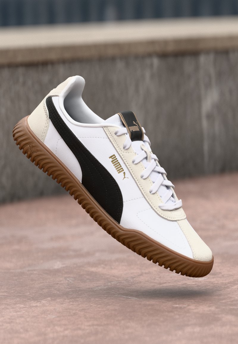 Hvide og sorte sneakers med gummisål, der har et struktureret sort stribe, guld logoaccent og klassisk snøredesign.