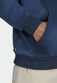 Námořnická modrá mikina s klokaní kapsou, žebrovanými manžetami a lemem. Hladká textura s jemným leskem, s malou logovou nášivkou na bočním švu.