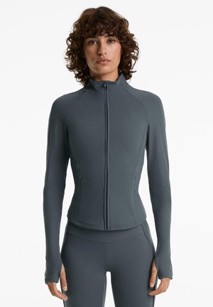 Vrouw in een strakke donkergrijze ritsjack met geribbelde lange mouwen en bijpassende hoge taille leggings, tegen een witte achtergrond.