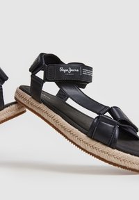 Musta sandaalit, joissa on sileä nahkapinta, säädettävät Velcro-hihnat ja kudottu juteemidsole. Kumipohja pitävän otteen takaamiseksi.