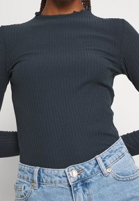 Top lungo grigio scuro a maniche lunghe con scollatura arricciata, abbinato a jeans di denim azzurri a vita alta con bottone metallico.