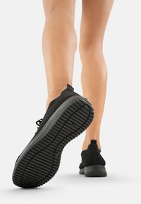 Zapatilla atlética negra con parte superior de punto texturizado y una suela de goma gruesa que presenta un patrón de labrado, vista de lado con un pie en movimiento.
