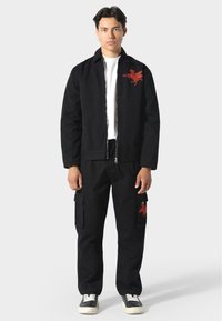 Schwarze Jacke mit Frontreißverschluss, weißes T-Shirt, schwarze Cargo-Hose mit Taschen und rotem Grafikdetail. Lässiges Design mit lockerer Passform.