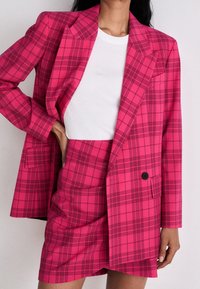 Růžový kostkovaný blazer s volným střihem a klopami, v kombinaci s odpovídající mini sukní. Tkanina má texturovaný pocit, černé knoflíkové detaily.
