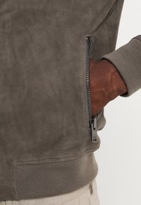 Giacca grigia in suede con polsini a costine, dotata di una tasca laterale con zip e una superficie liscia. Il tessuto ha una finitura opaca.
