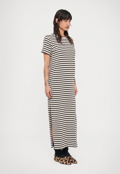 Γυναίκα που φοράει μακρύ φόρεμα με καφέ και λευκές οριζόντιες ρίγες και κοντά μανίκια, μαύρες κάλτσες και παπούτσια με λεοπάρ μοτίβο, στέκεται πλάγια.