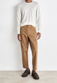 Maglione a maniche lunghe di un beige chiaro, pantaloni cargo color cammello con tasche laterali, e mocassini marroni con suola spessa. Stile semplice, vestibilità rilassata e texture minima.