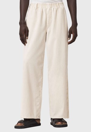 Osoba ubrana w luźne beżowe spodnie sztruksowe z elastycznym pasem i czarne sandały, stojąca na jednolitym tle.