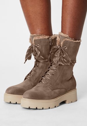 Bruine suède enkellaarsjes met dikke beige zolen, gevoerd met nepbont, voorveters en een zijrits, gedragen door donkergekleurde benen.