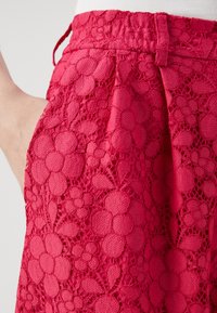I pantaloni corti rossi in pizzo presentano un motivo floreale, una superficie testurizzata e una vita elastica con cordoncino. Tasca laterale visibile a sinistra.