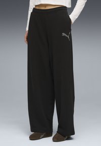 Άτομο με χαλαρό μαύρο παντελόνι φόρμας Puma με λογότυπο από στρας, λευκό κοντό μπλουζάκι με μακριά μανίκια και καφέ παπούτσια χωρίς κορδόνια, στέκεται ακίνητο.