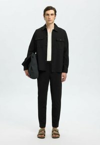 Schwarzes Hemd mit Knöpfen über einem hellen Hemd, kombiniert mit schwarzen Hosen und olivgrünen Sandalen. Hält eine graue Tasche mit schickem Design.